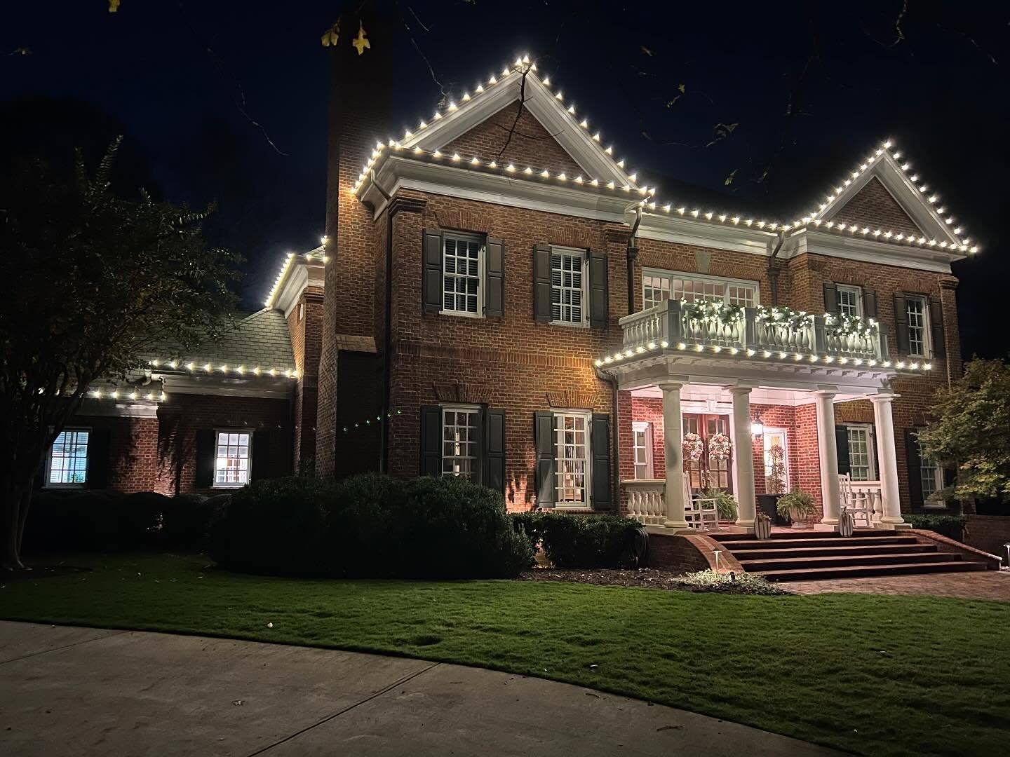 Red-brick Georgian colonial with roofline lights and lit balcony greenery over a columned porch