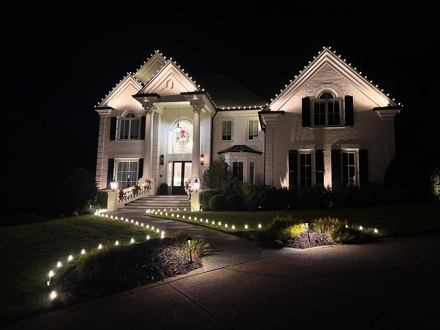 Grand white-painted brick estate with lit roofline peaks and garland with red bows on the porch rail