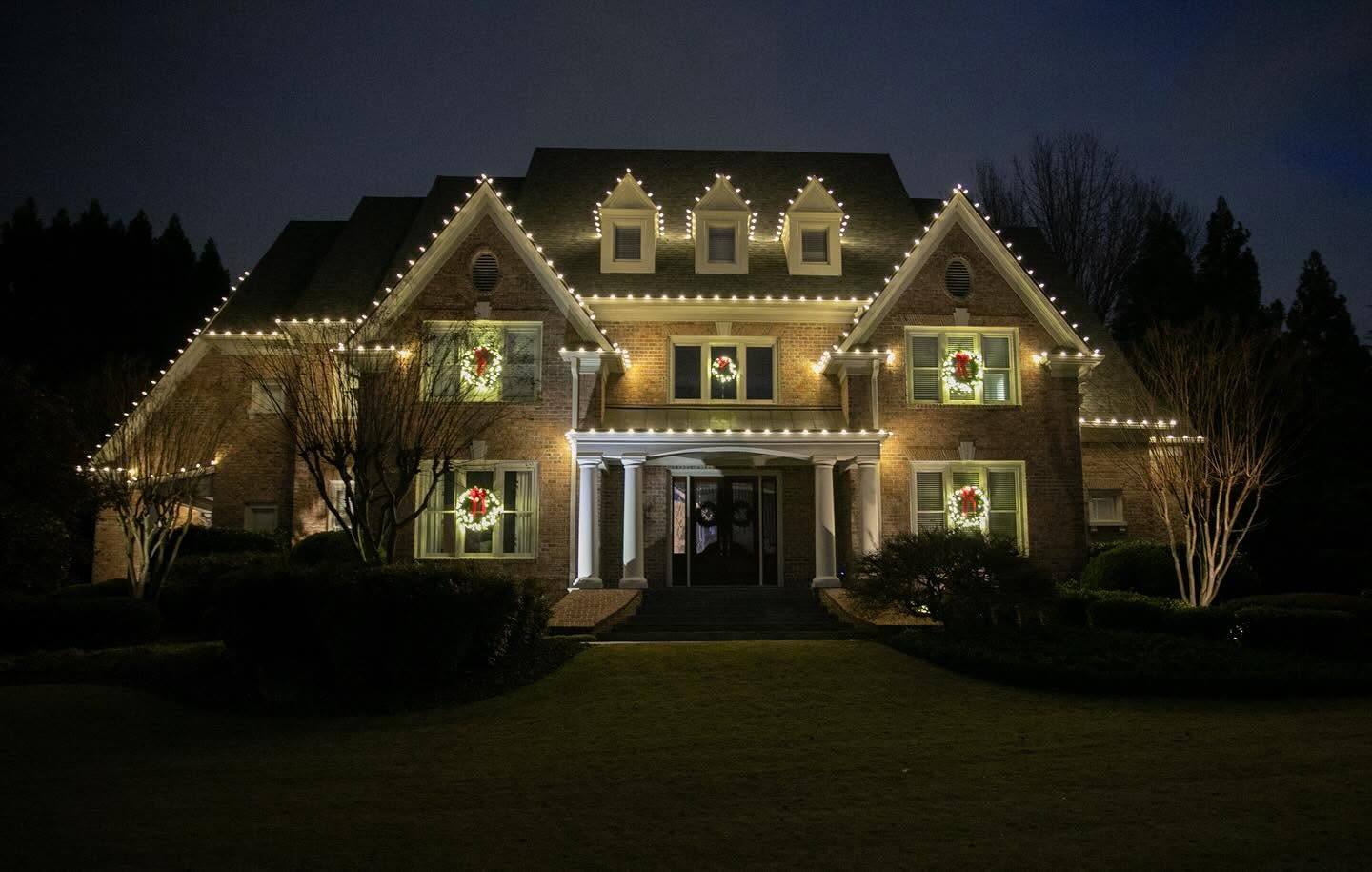 Large brick Tudor-style estate with a lit roofline and wreaths in the windows